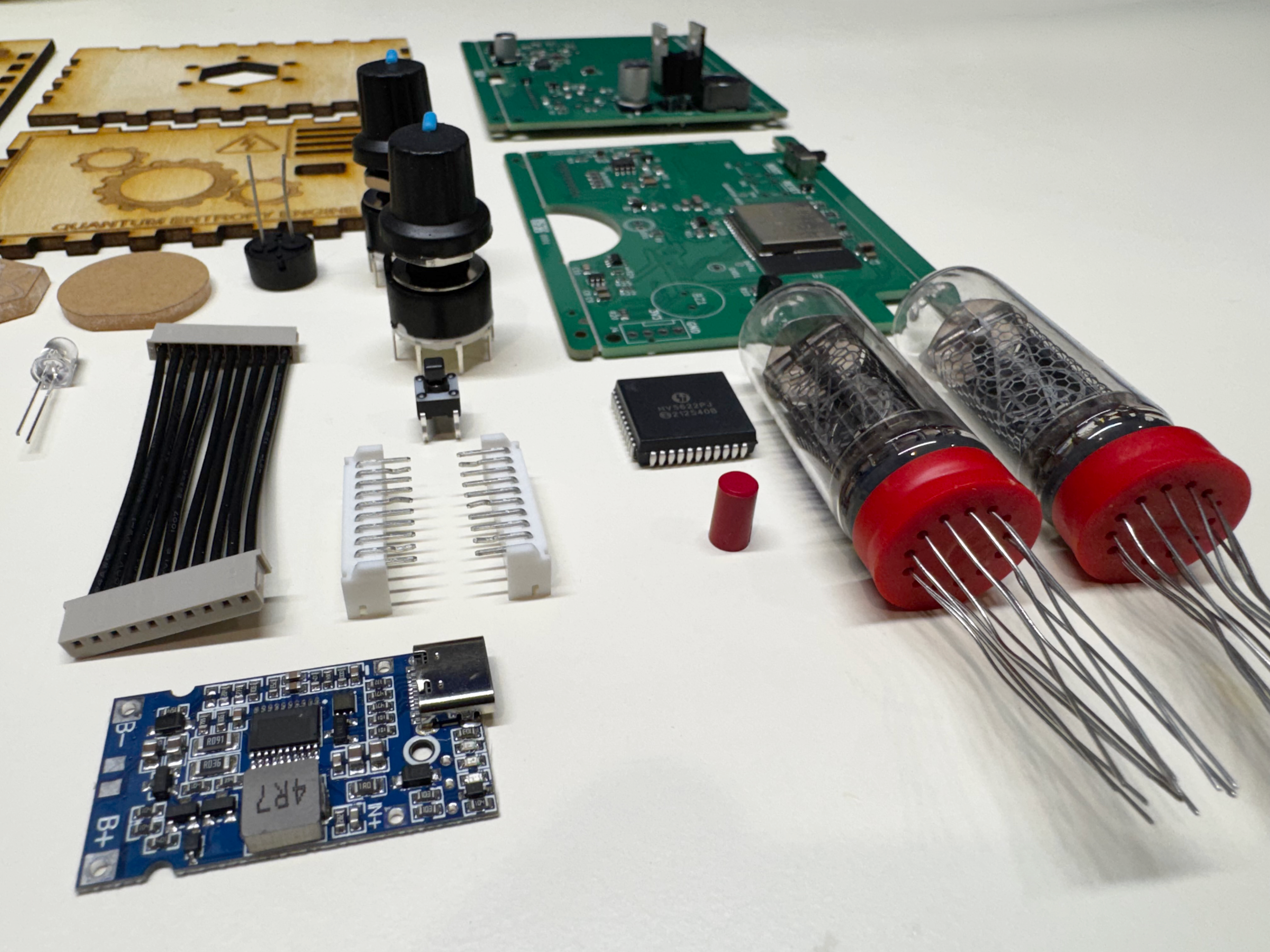 Components of a disassembled quantum entropy engine: expedition kit. The components are on a white laboratory bench. The components in the view include two circuit boards, some electrical components (rotary knobs, board to board cable harness, JST connectors, a high voltage display IC), the charging PCBA, and the Nixie tubes.